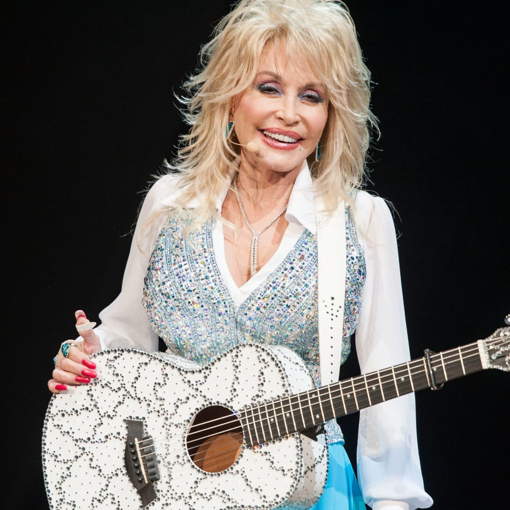 Dolly Parton holding an acoustic guitar on stage in a white bedazzled outfit.