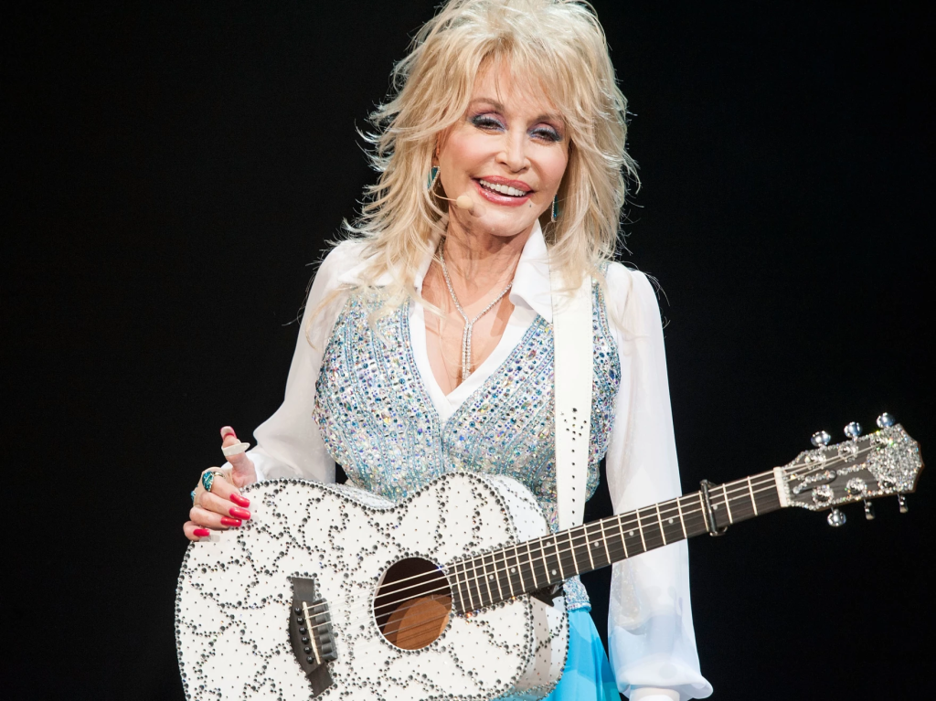 Dolly Parton holding an acoustic guitar on stage in a white bedazzled outfit.