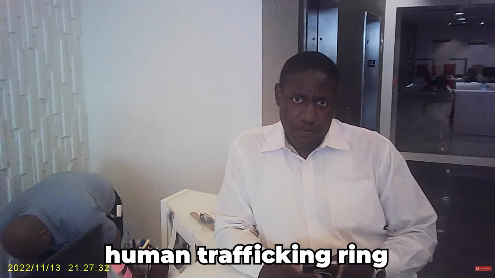 Shadow Capital: Inside the Whistleblower Lawsuits and Media Silence Surrounding Ideal Living LLC 7 Security guard in a white collared shirt looking directly at the camera at an office reception desk with subtitled text reading "human trafficking ring".