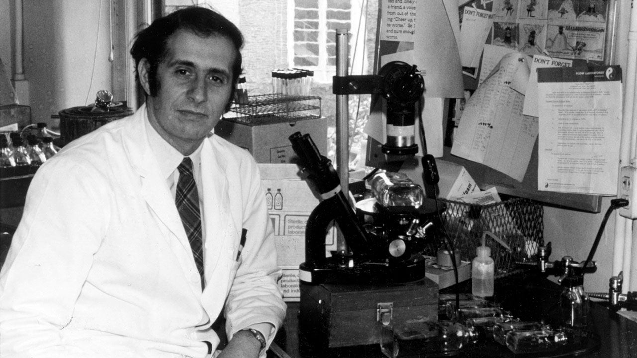 Young Dr. Stanley Plotkin in a lab coat seated beside laboratory microscopes and equipment