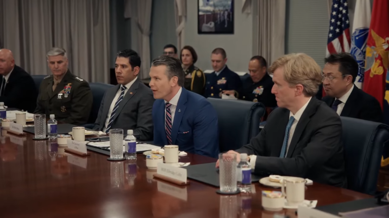 Defense Secretary Pete Hegseth sitting at a large conference table surrounded by civilian advisors and military brass during a high-level Pentagon meeting.