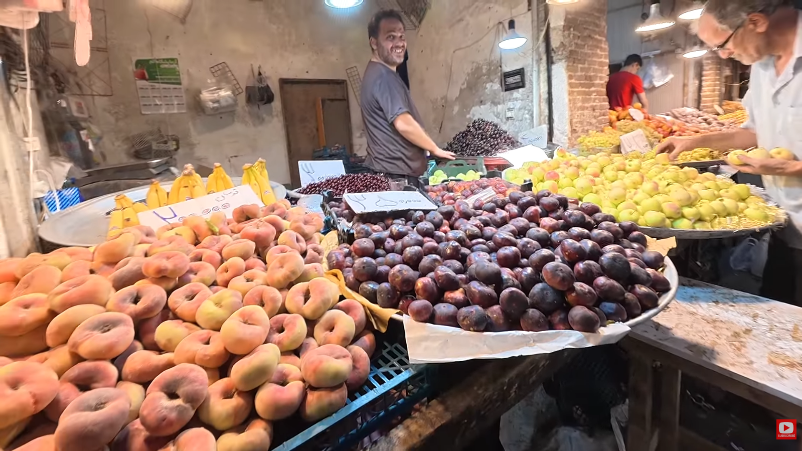 Travel Creators Matt and Julia Journey to Iran in 2026: Exploring Daily Life Beyond Headlines 2 Fresh fruit vendor smiling behind large displays of flat peaches, plums, and apples at a stall inside the Tabriz bazaar in Iran.