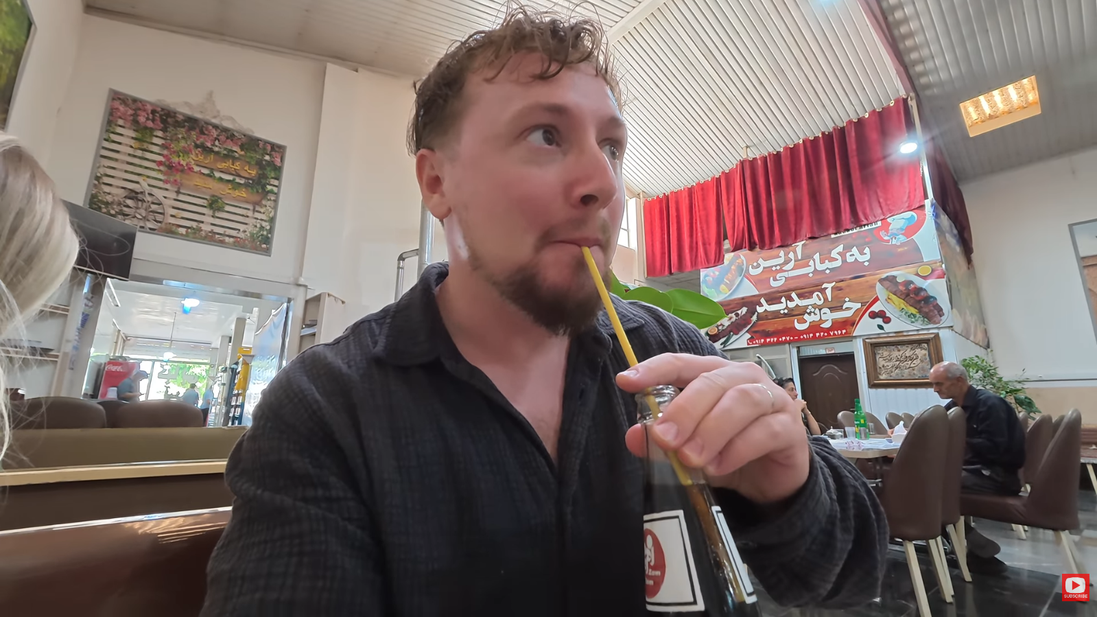 Travel Creators Matt and Julia Journey to Iran in 2026: Exploring Daily Life Beyond Headlines 3 Travel vlogger Matt drinking a glass bottle of Zam Zam cola through a straw while sitting inside a local restaurant in Bonab, Iran.