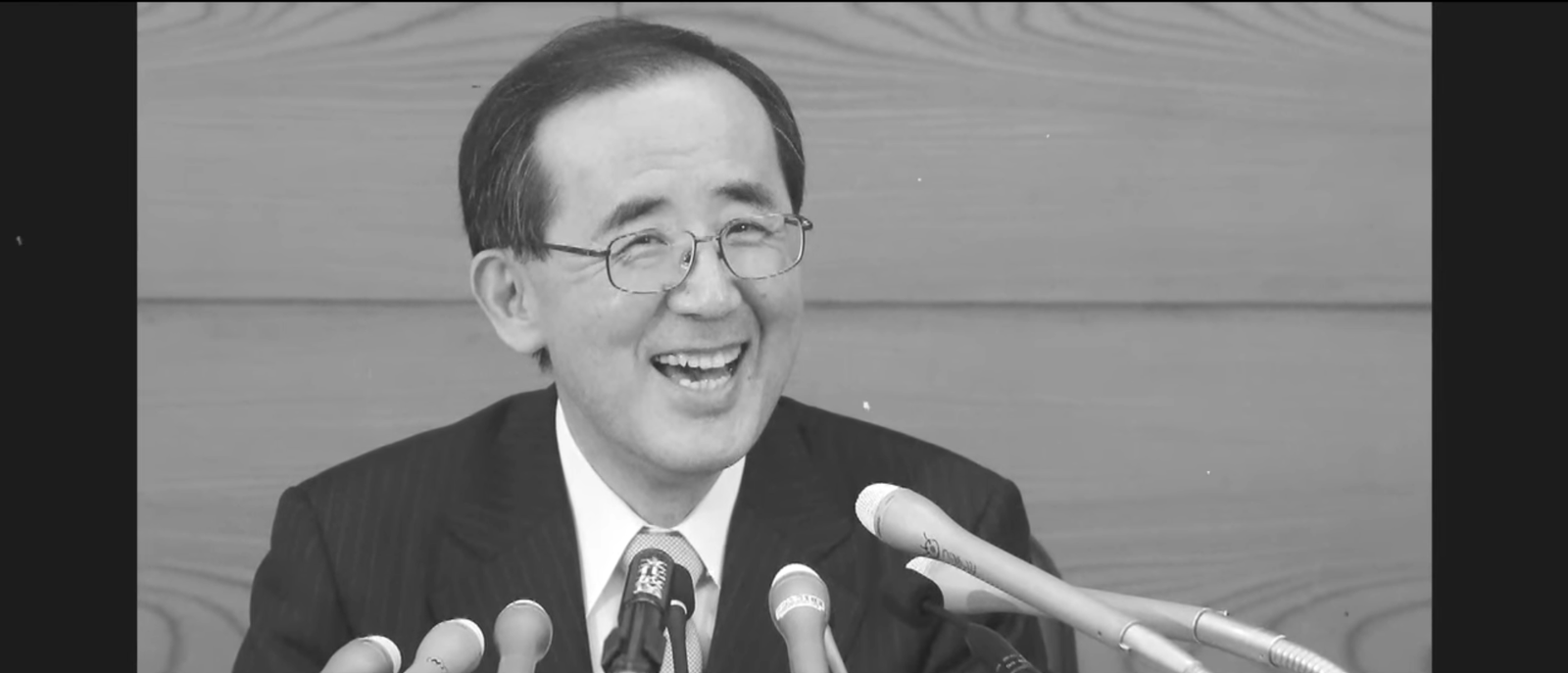 Black and white photograph of former Bank of Japan Governor Masaaki Shirakawa smiling at a press conference microphone array