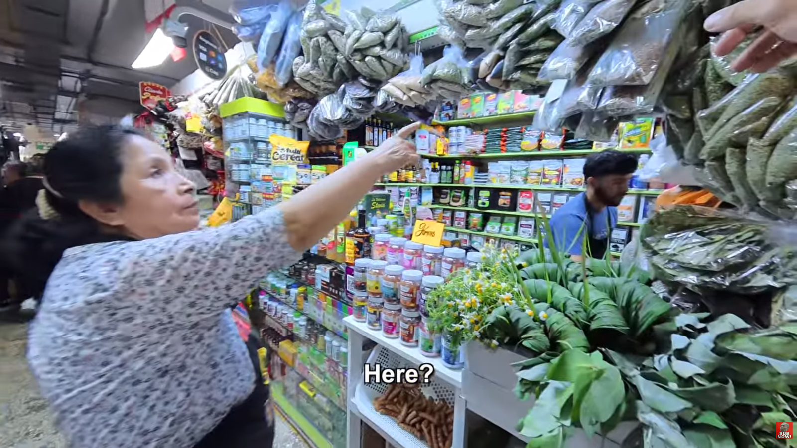 A local female market vendor pointing toward bags of natural cocoa leaves and herbal remedies for Kurt Caz at a stall in Mercado Central, Lima.