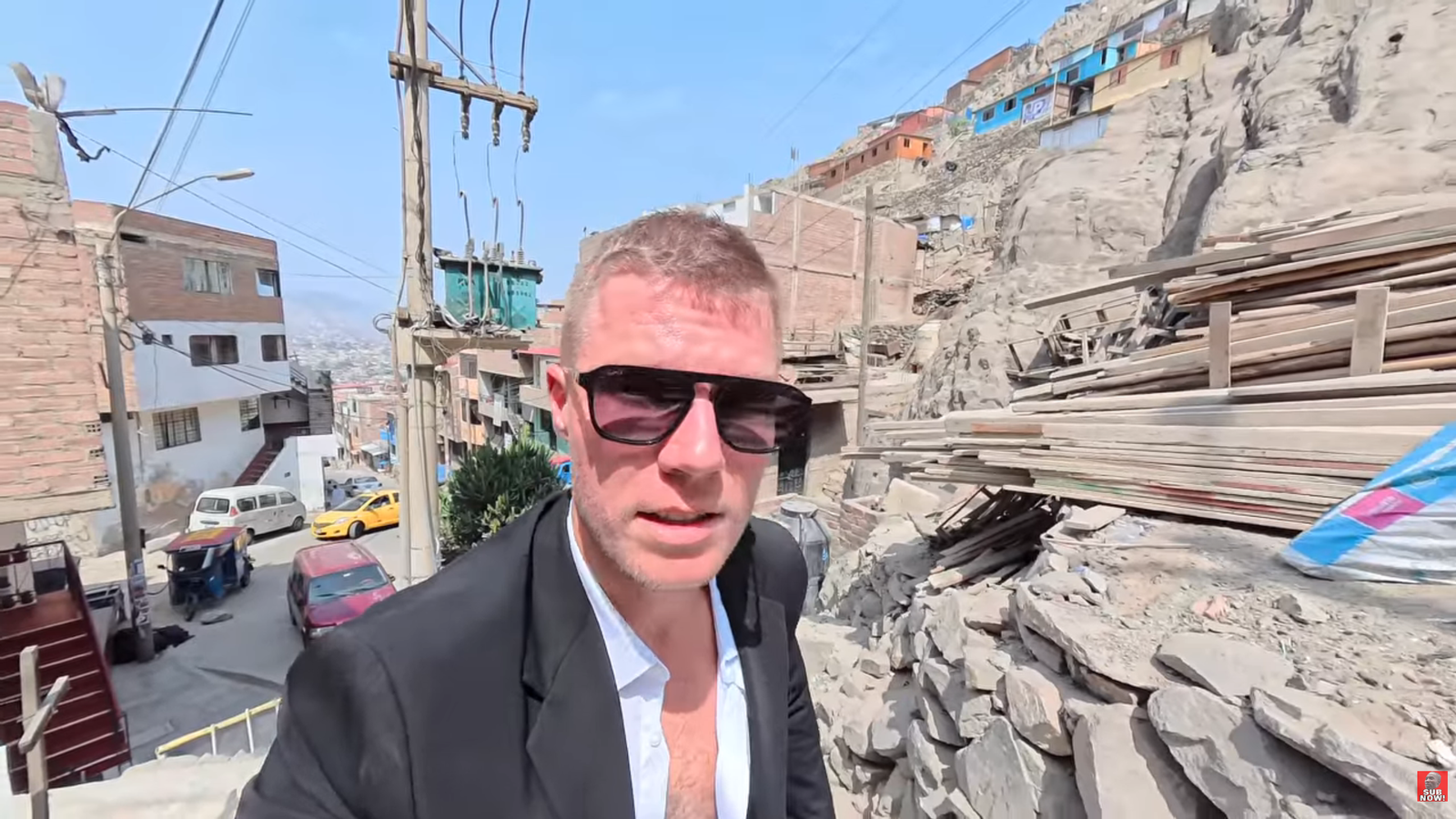 Travel vlogger Kurt Caz wearing sunglasses and a blazer while filming in the steep hillside neighborhood of San Juan de Lurigancho in Lima, Peru.