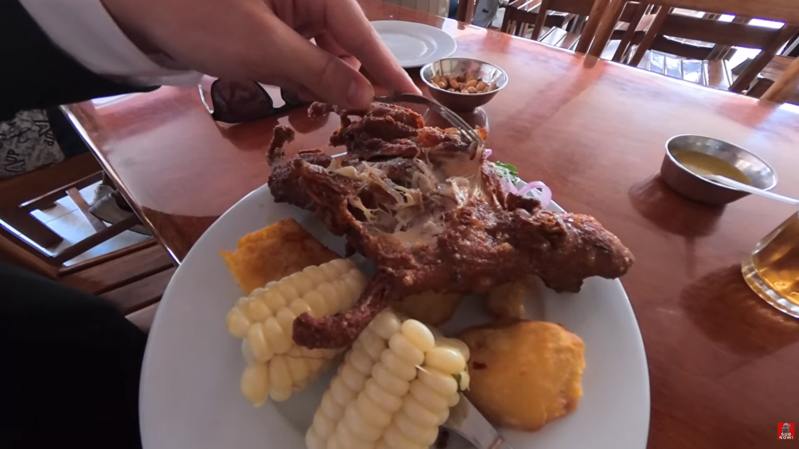 Close-up of a plate of traditional Peruvian Cuy Chactado (fried guinea pig) served with giant Andean corn and potatoes, with Kurt Caz holding a piece.