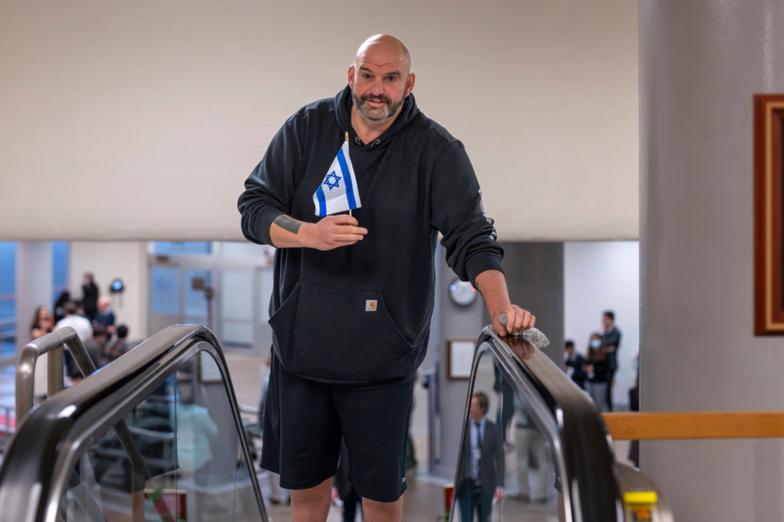 Senator John Fetterman (D-PA) in a black Carhartt hoodie, holding a small Israeli flag on an escalator.